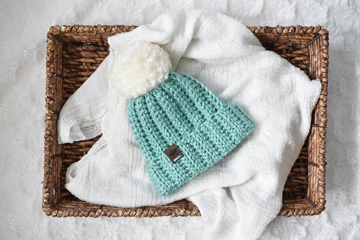 A green crochet beanie with a fluffy white pom pom sits on a wooden basket and white sheet. Easy Breezy Chunky Beanie crochet pattern by The Turtle Trunk.