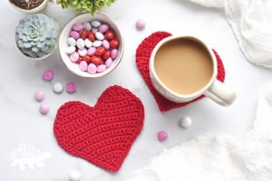 2 red crochet heart coasters, one with a coffee cup on it. bowl of candy in the background.