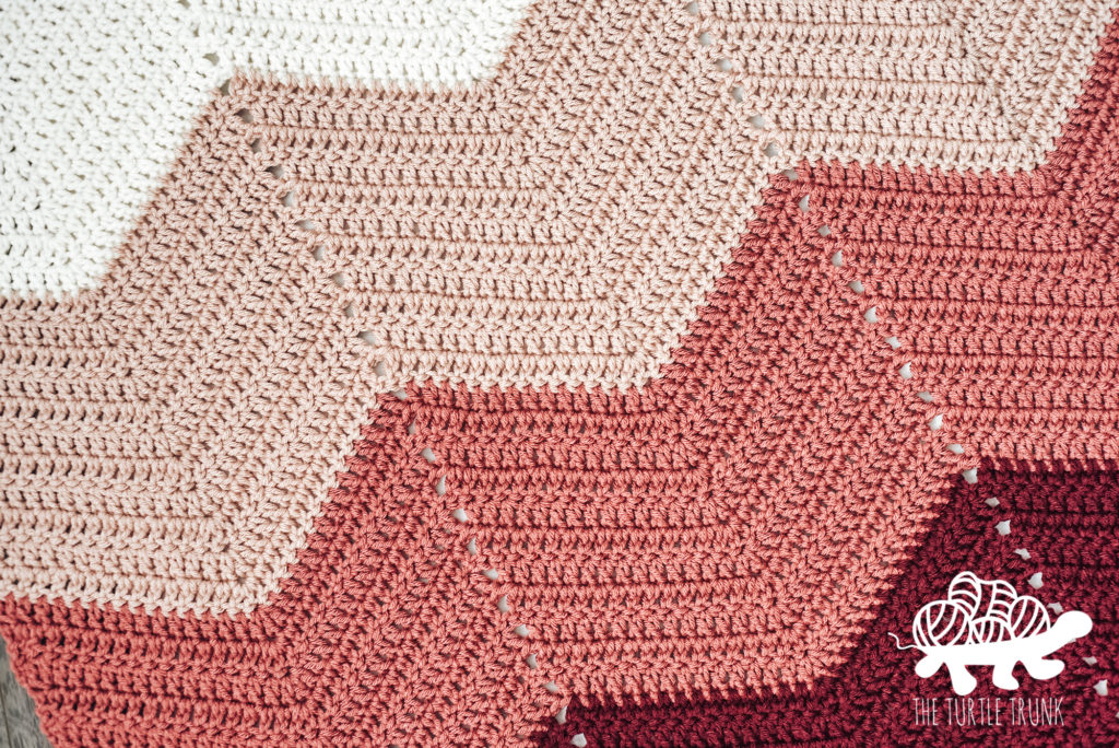 Close up photo of a pink, ombre, chevron crochet blanket laying on the ground. Bebe Baby Blanket crochet pattern by The Turtle Trunk