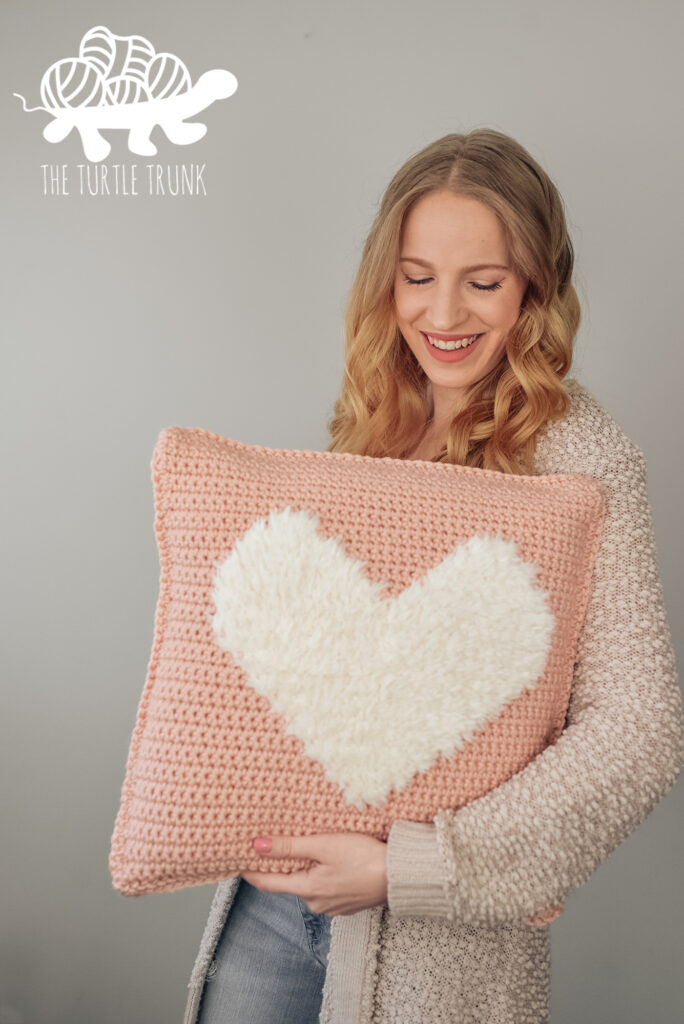 Photo shows a woman holding up a crochet pillow that features a faux fur heart in the center. Crochet pattern is the Bulky Love to Cuddle Pillow by The Turtle Trunk.