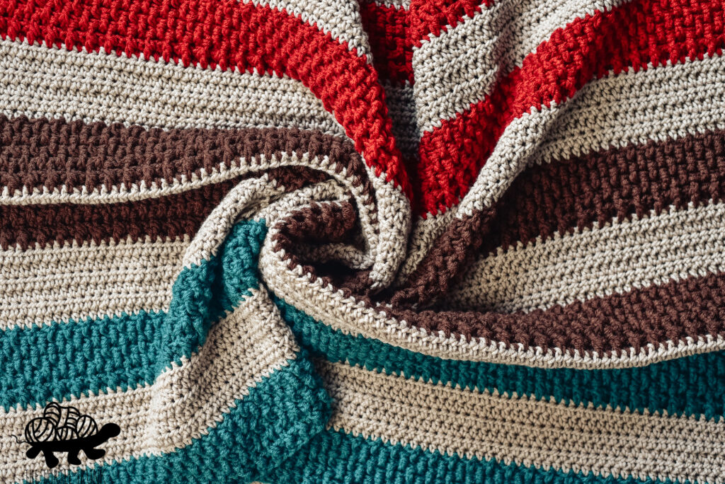 Photo shows a red, beige, brown, and teal crochet blanket laying on the ground. Crochet pattern is the Bonfire Blanket by The Turtle Trunk.