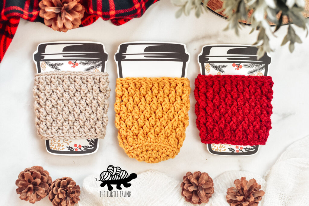 Photo shows 3 crochet cup cozy's laying on a white surface. Crochet pattern is the Country Cottage Cozy by The Turtle Trunk.