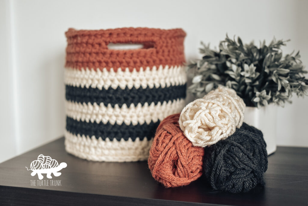 Striped crochet basket sitting on a desk beside a plant and with 3 skeins of yarn sitting in front.