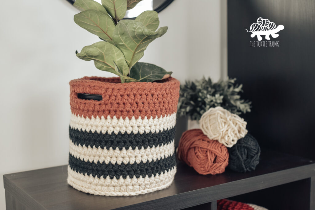 Brecken Basket (crochet basket) holding a fiddle leaf fig plant on a shelf. Behind the basket sits 3 skeins of yarn and another plant.