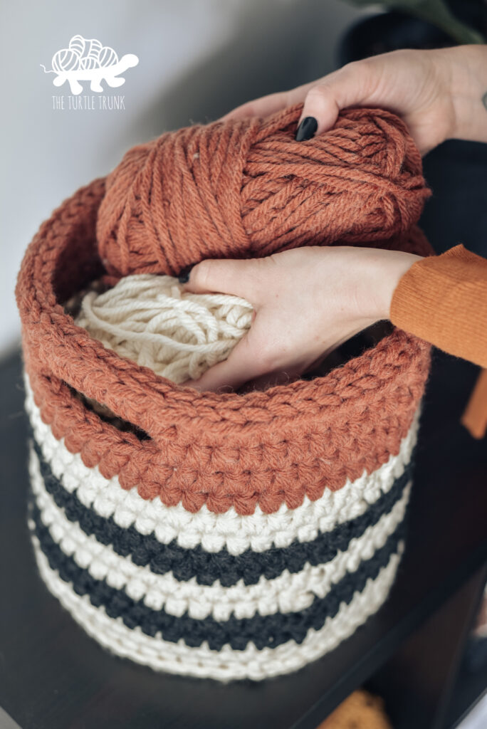 Striped crochet basket sitting on a desk with hands putting yarn inside the basket.