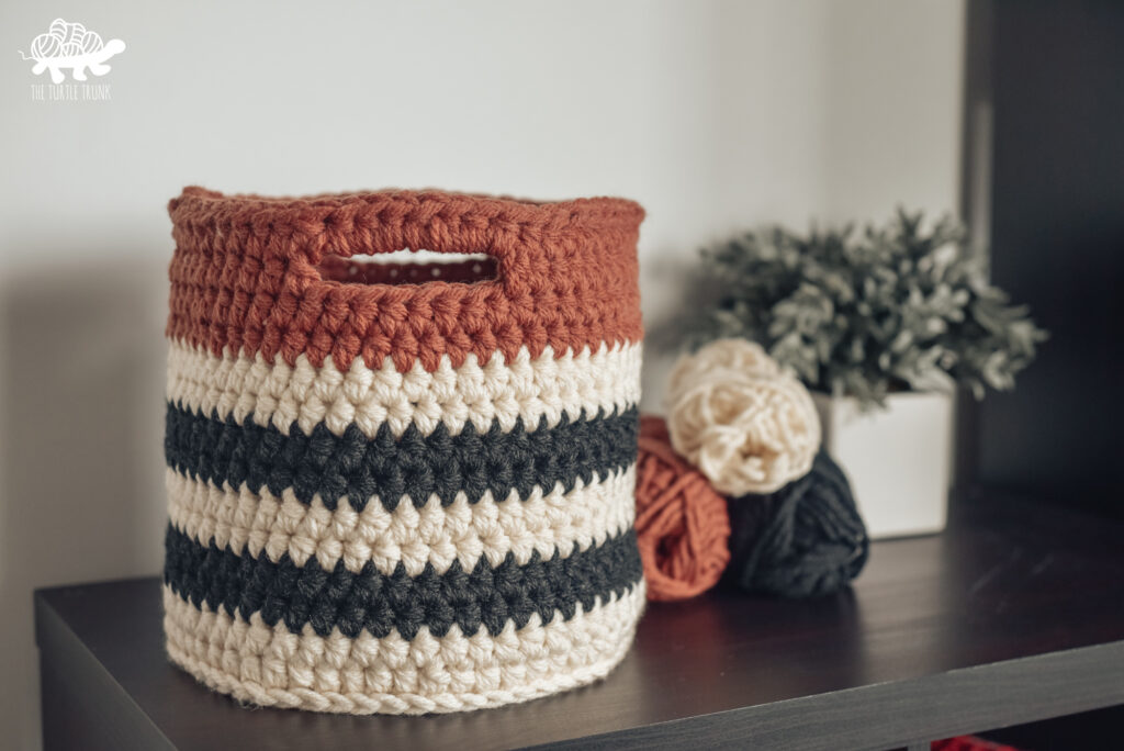 Orange, black, and white striped crochet basket sitting on a table with yarn and plant behind it. 