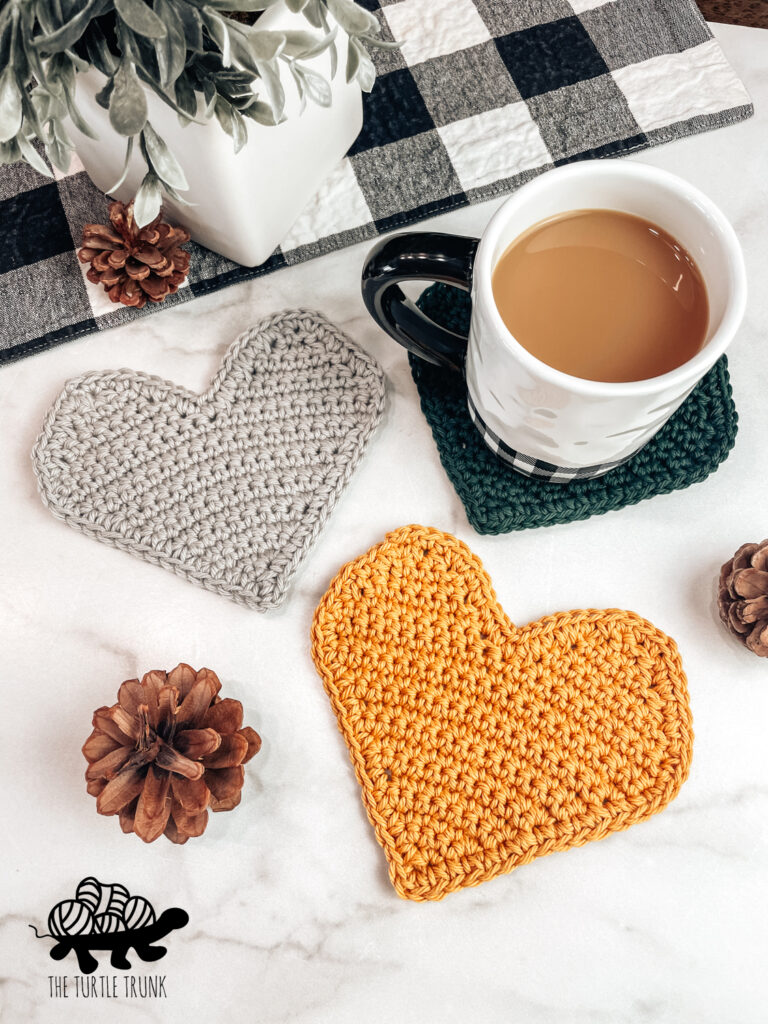 3 crochet heart shaped coasters laying on white background, one with a coffee mug on it.