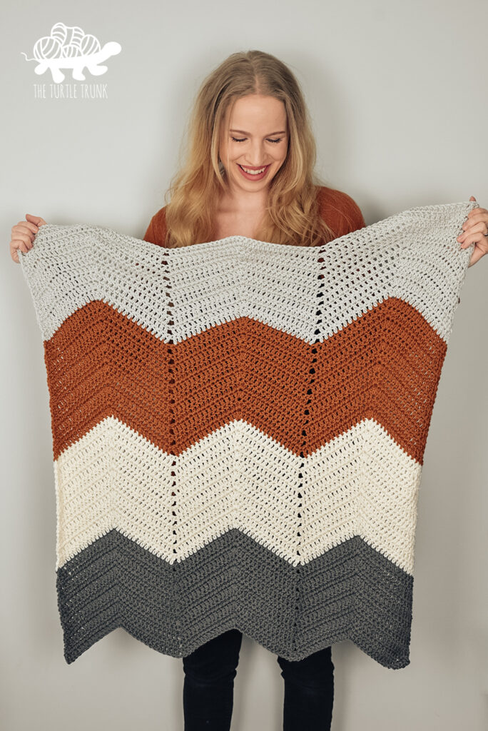 Woman holding a Bebe Baby Blanket in the colors light grey, burnt orange, white, and dark grey.