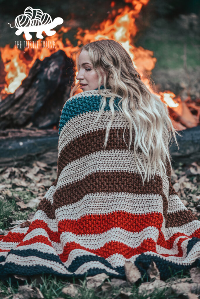 Women sitting outside by a fire, with a striped, crochet blanket wrapped around her shoulders. Crochet blanket is the Bonfire Blanket crochet pattern by The Turtle Trunk