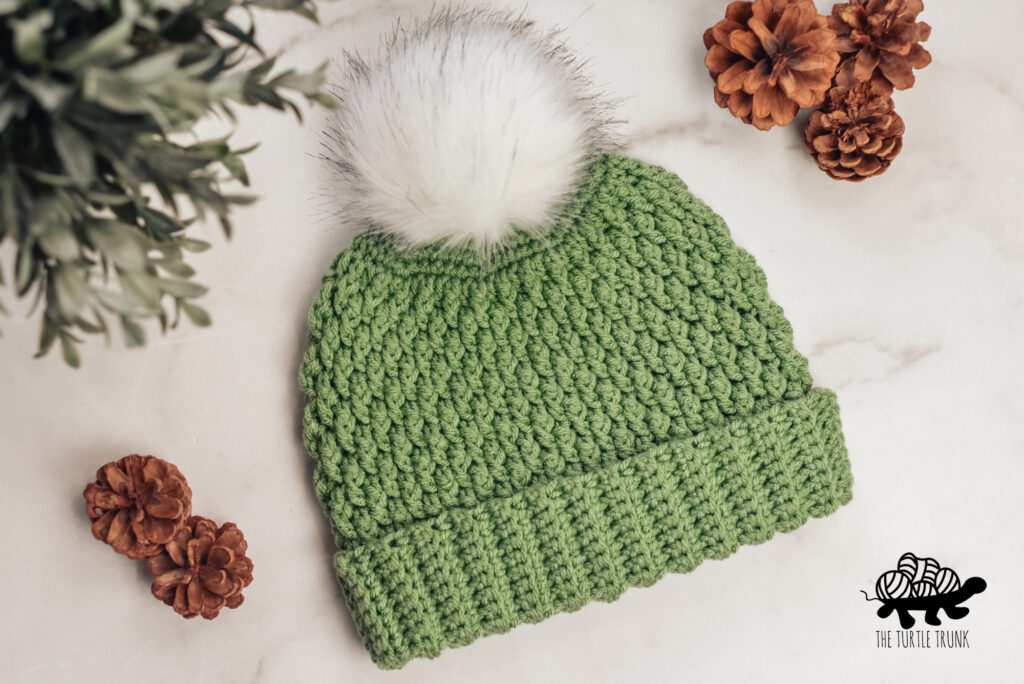 A green, crochet beanie with a. white faux fur pom pom attached, lays on a white surface. Country Cottage Beanie crochet pattern by The Turtle Trunk.