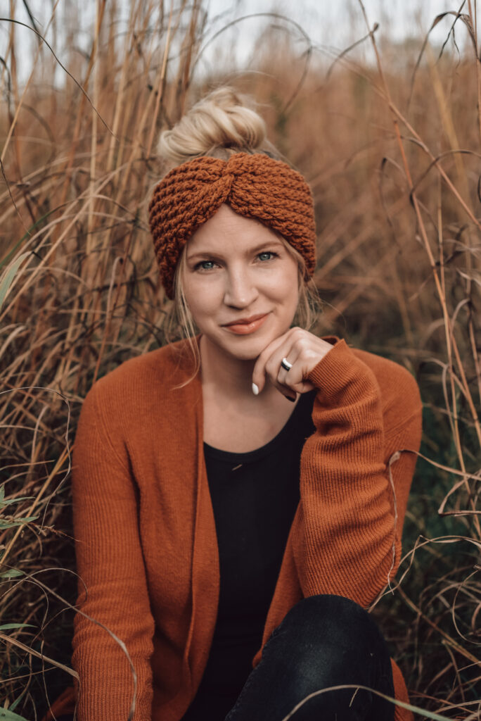 Photo shows a photo of a woman wearing a orange crochet headband, orange sweater, and black tank top. Photo is of Ali Wholihan, the crochet designer behind The Turtle Trunk.