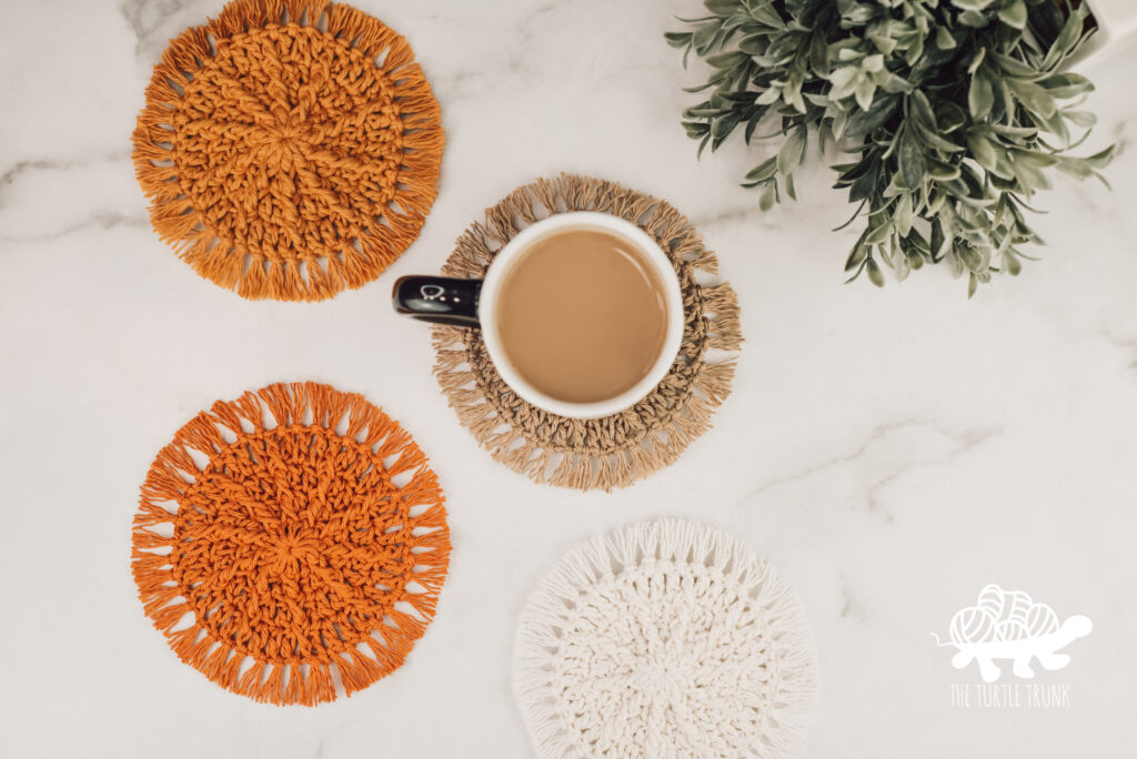 Photo shows 4 crochet coasters laying on a white surface. A cup of coffee is sitting on one of the coasters. Coaster is the Hello Sunshine Coaster crochet pattern by The Turtle Trunk.