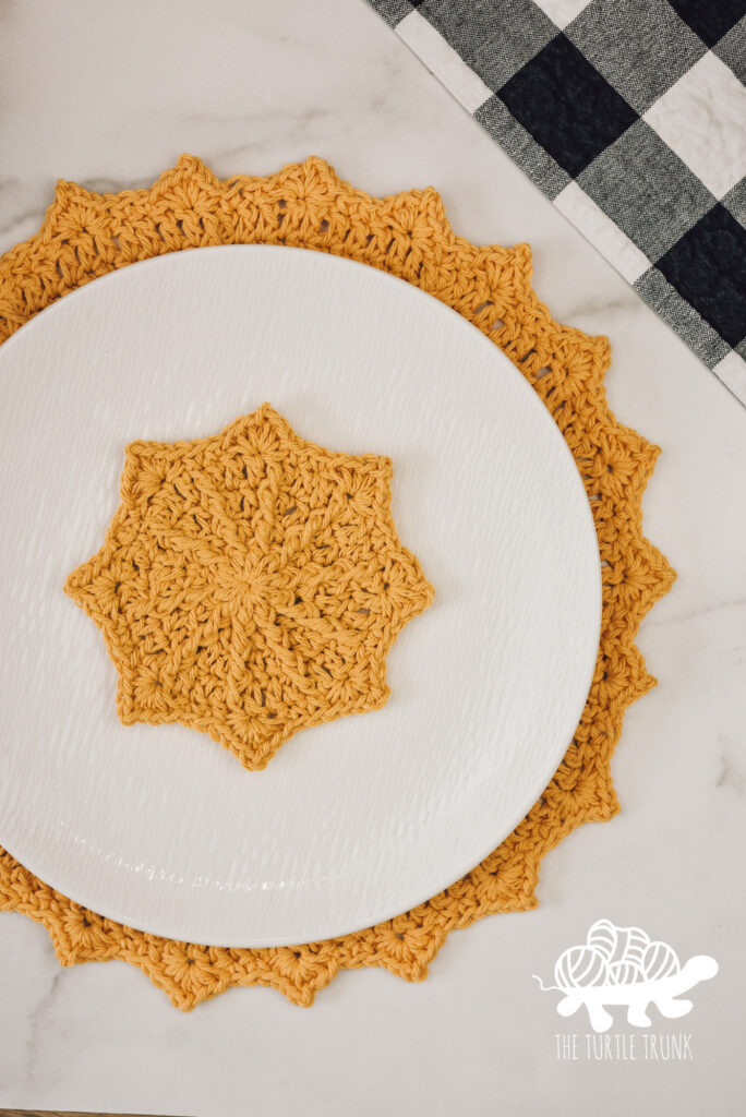 Photo shows a yellow, crochet coaster and placemat laying on a white surface. Crochet patterns are the Hello Sunshine Placemat and Hello Sunshine Coaster by The Turtle Trunk.