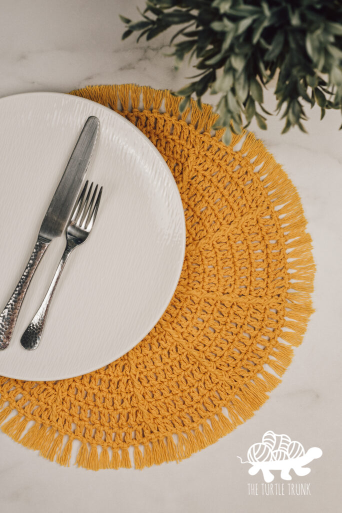 Photo shows a round, yellow, crochet placemat laying on a white surface. The placemat has a white plate and fork and knife on it.