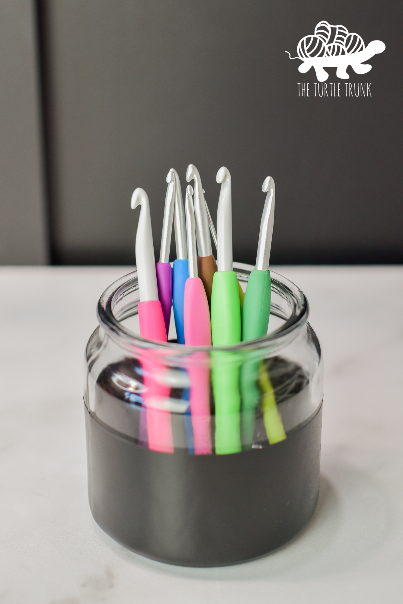 Photo of crochet hooks in a glass jar sitting on a white surface and black background.