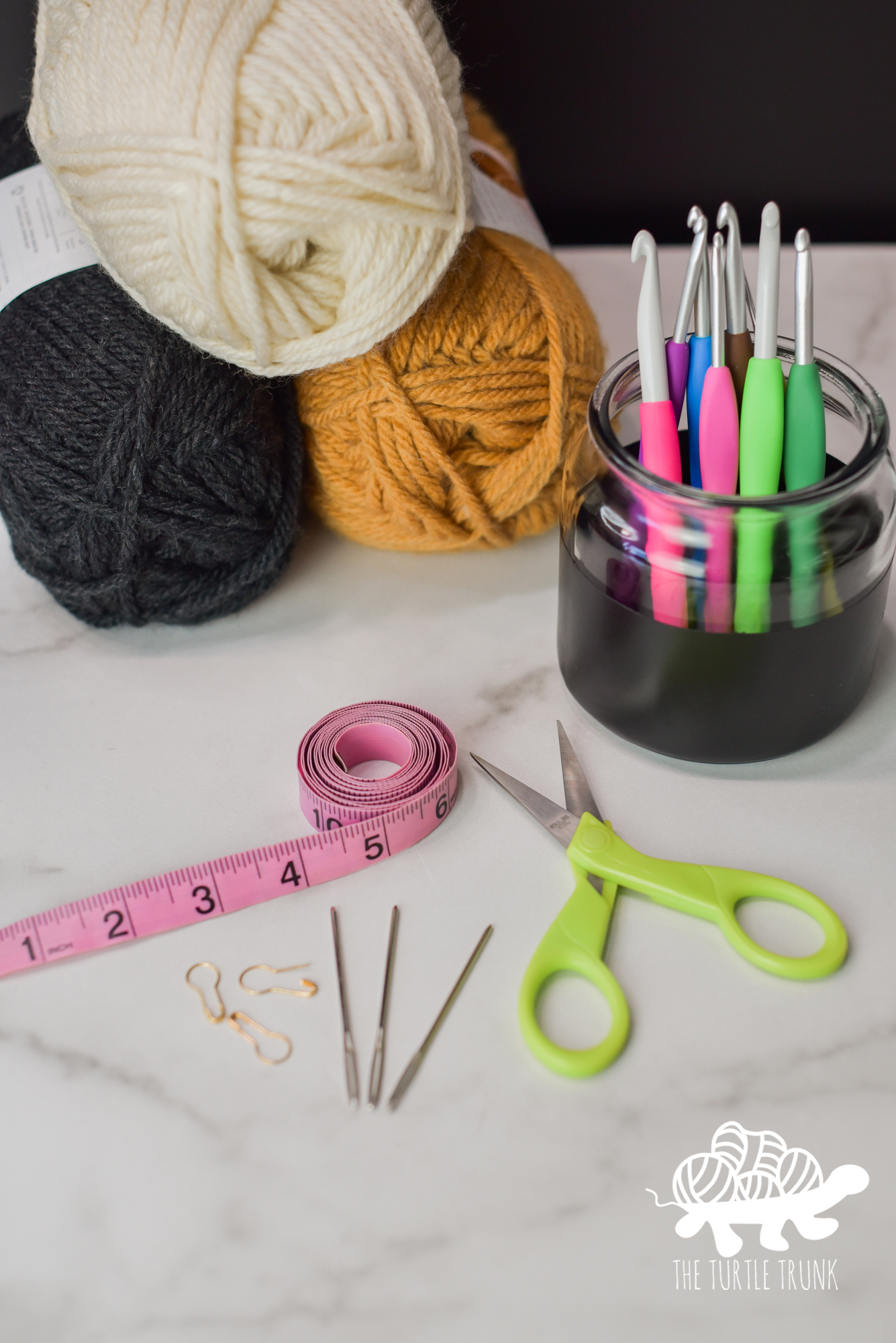 Photo of supplies needed for crochet, such yarn 3 skeins of yarn, crochet hooks, yarn needles, stitch markers, a measuring tape, and scissors lay on a white surface.