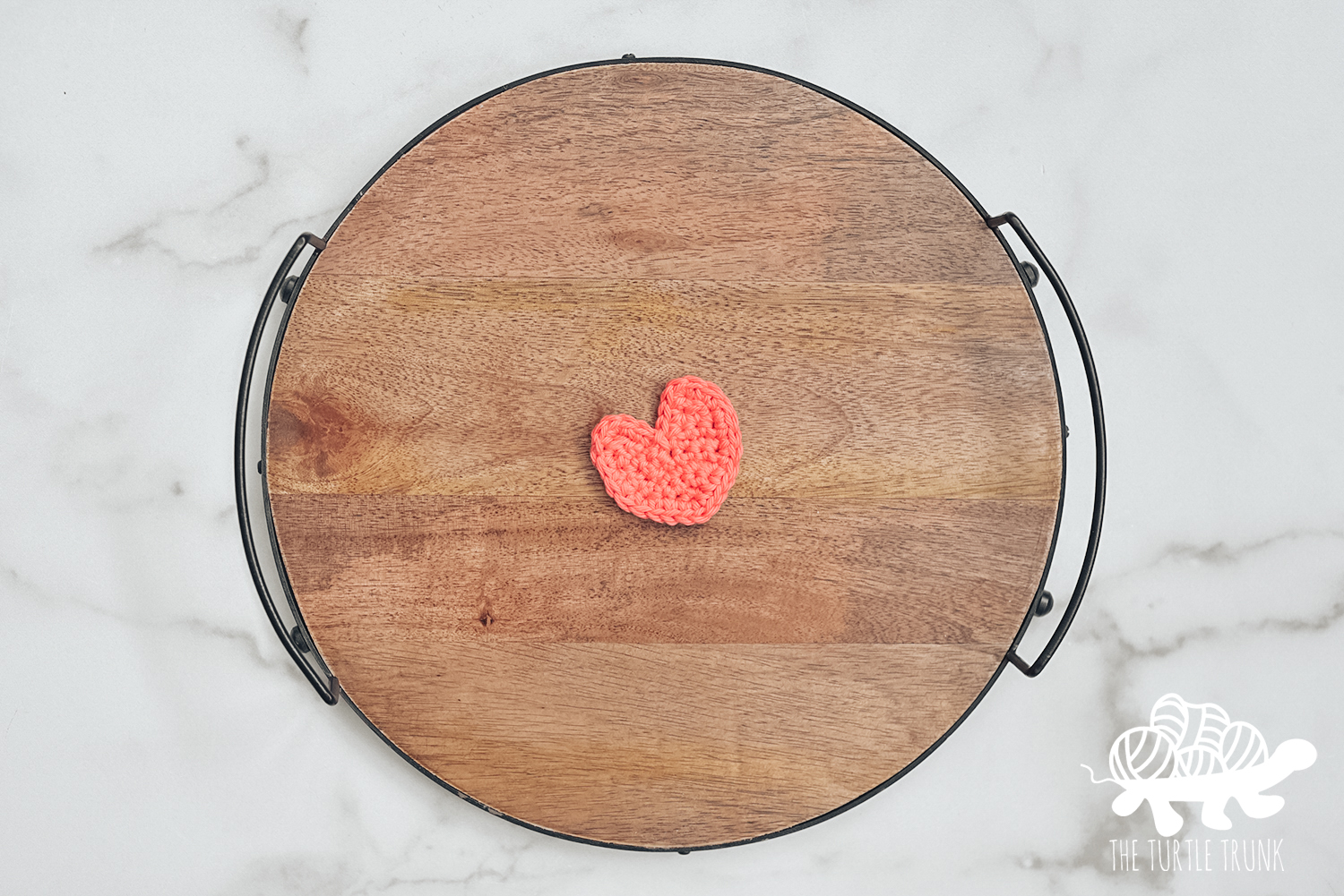 A mini crochet heart sits on a round, wooden board on a white surface.