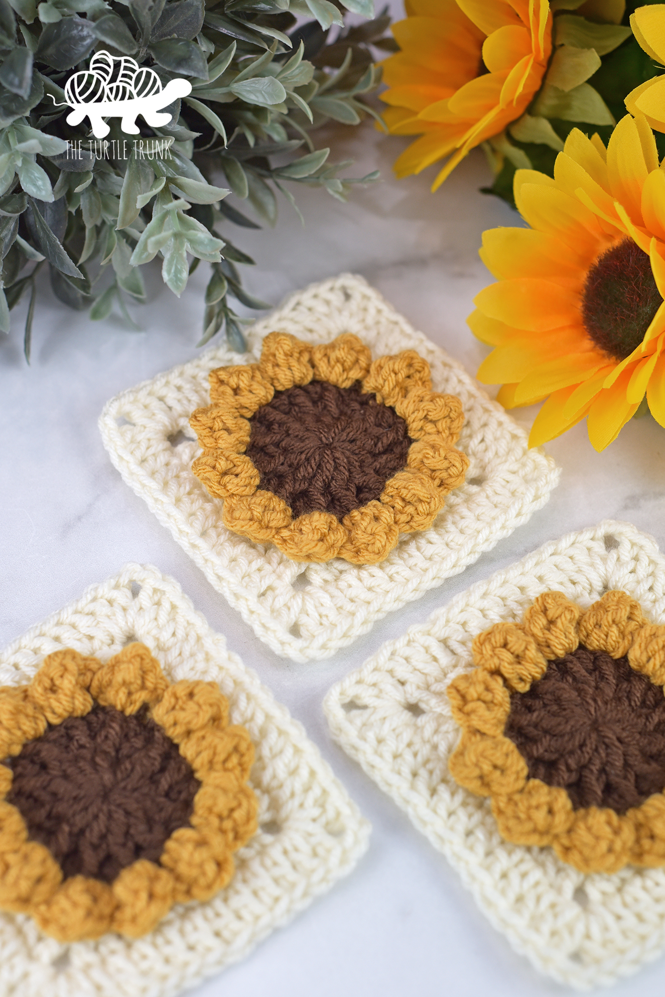 3 crochet sunflower squares lay on a white surface with real real sunflowers and plants surrounding them.