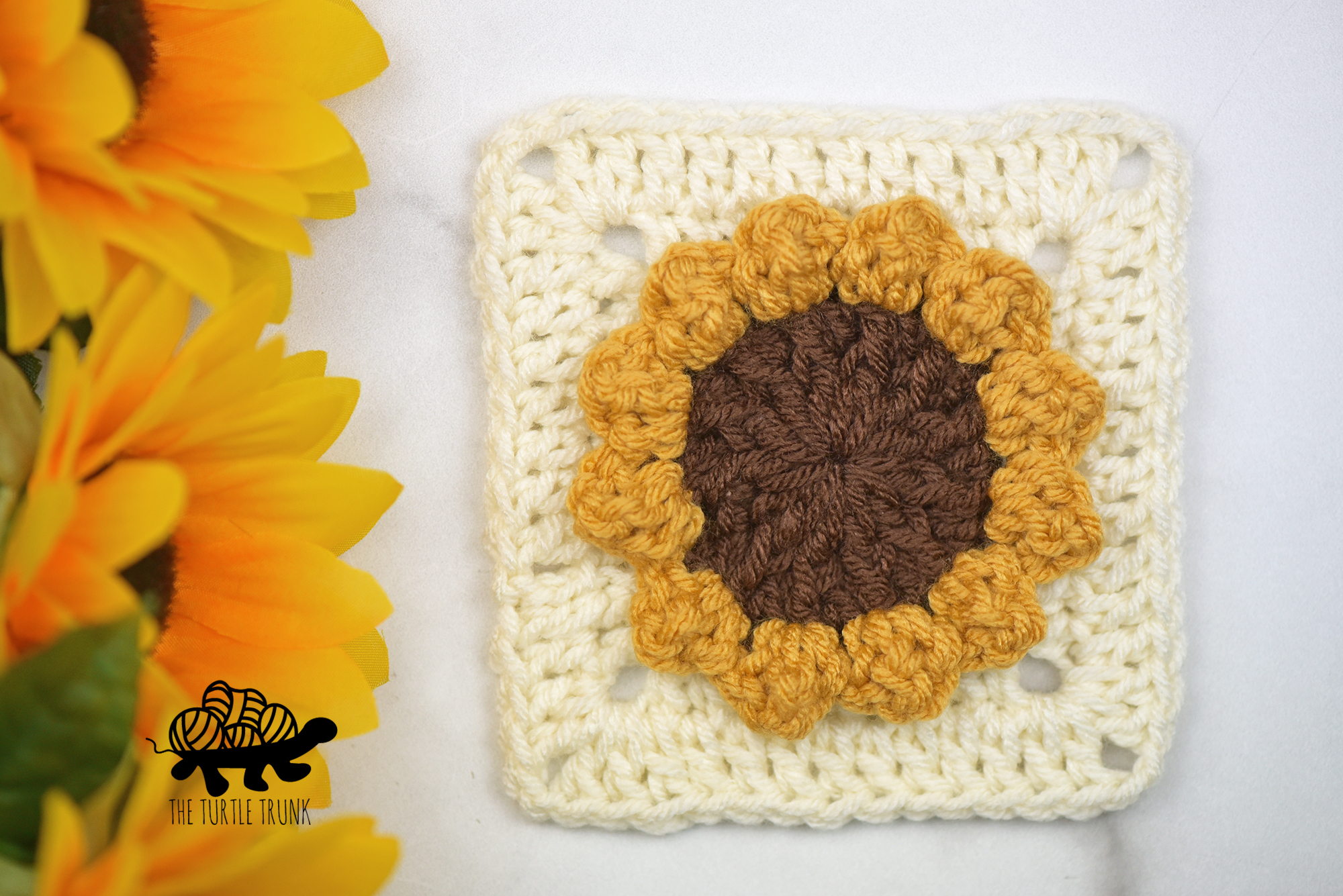 A crochet sunflower square laying on a white surface with real sunflowers beside.