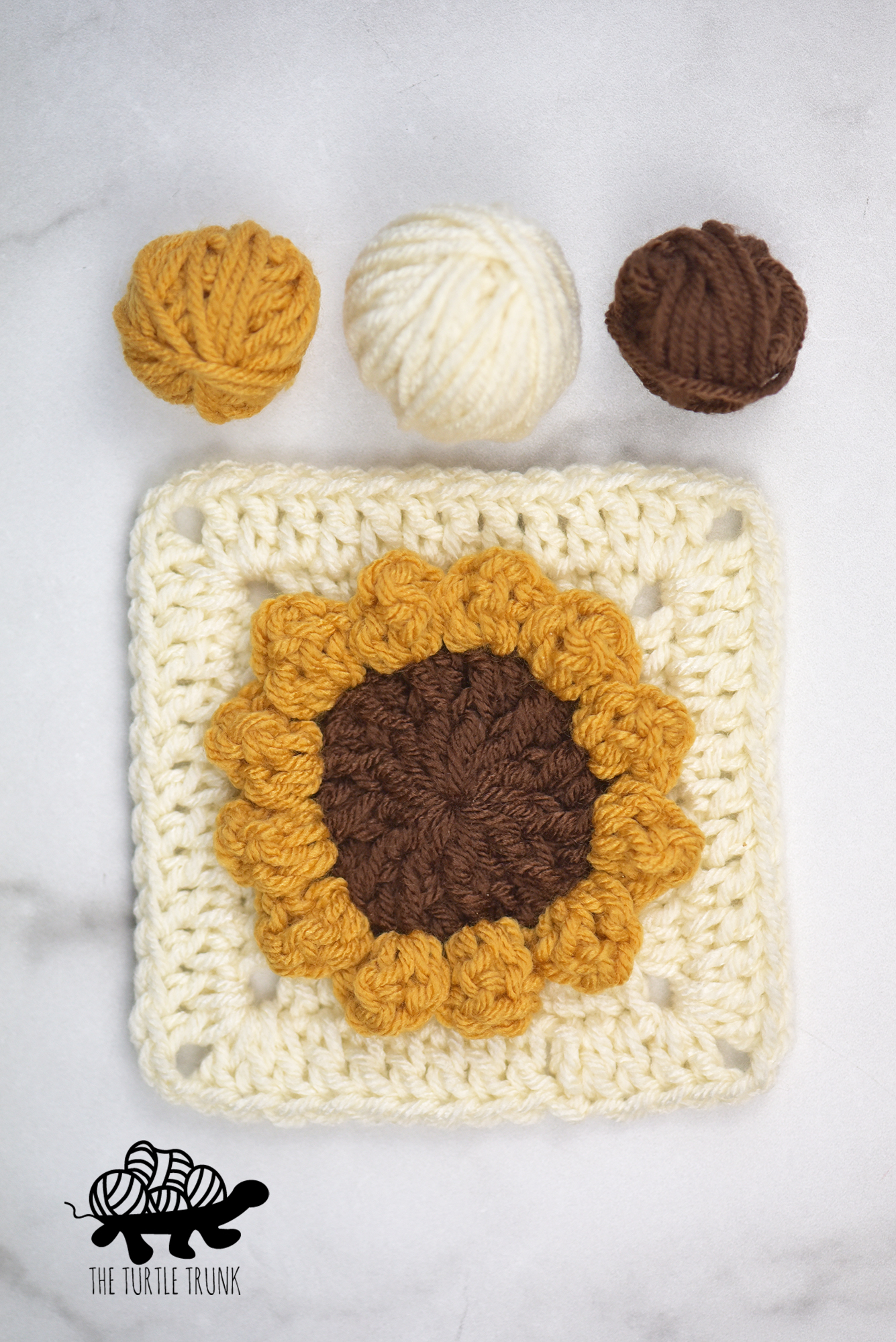 3 balls of yarn and a crochet sunflower square lay on a white surface. Sunflower Square crochet pattern by The Turtle Trunk.