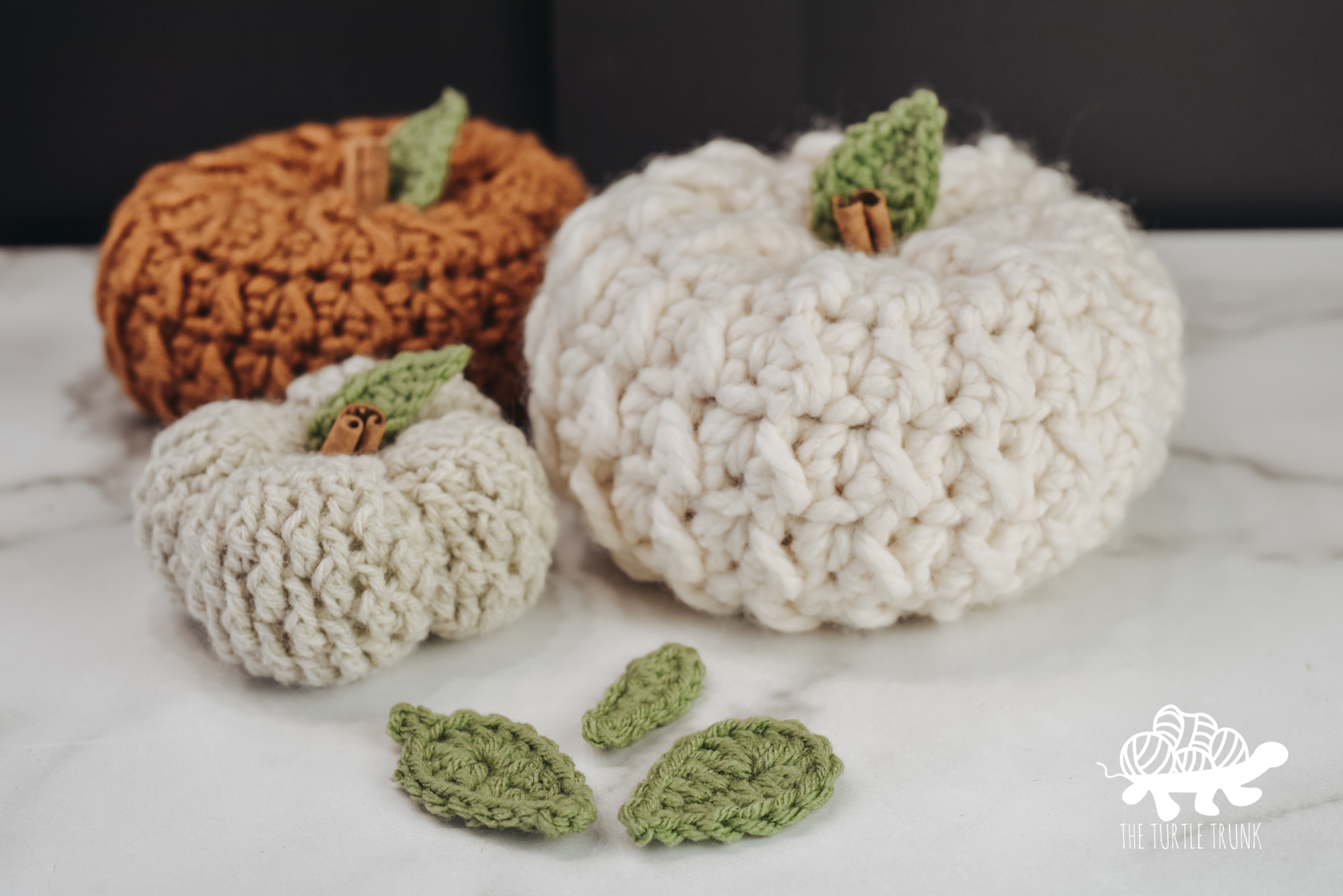 Photo shows 3 crochet pumpkins with crochet leaves in top. 3 crochet leaves sit in front of the pumpkins.