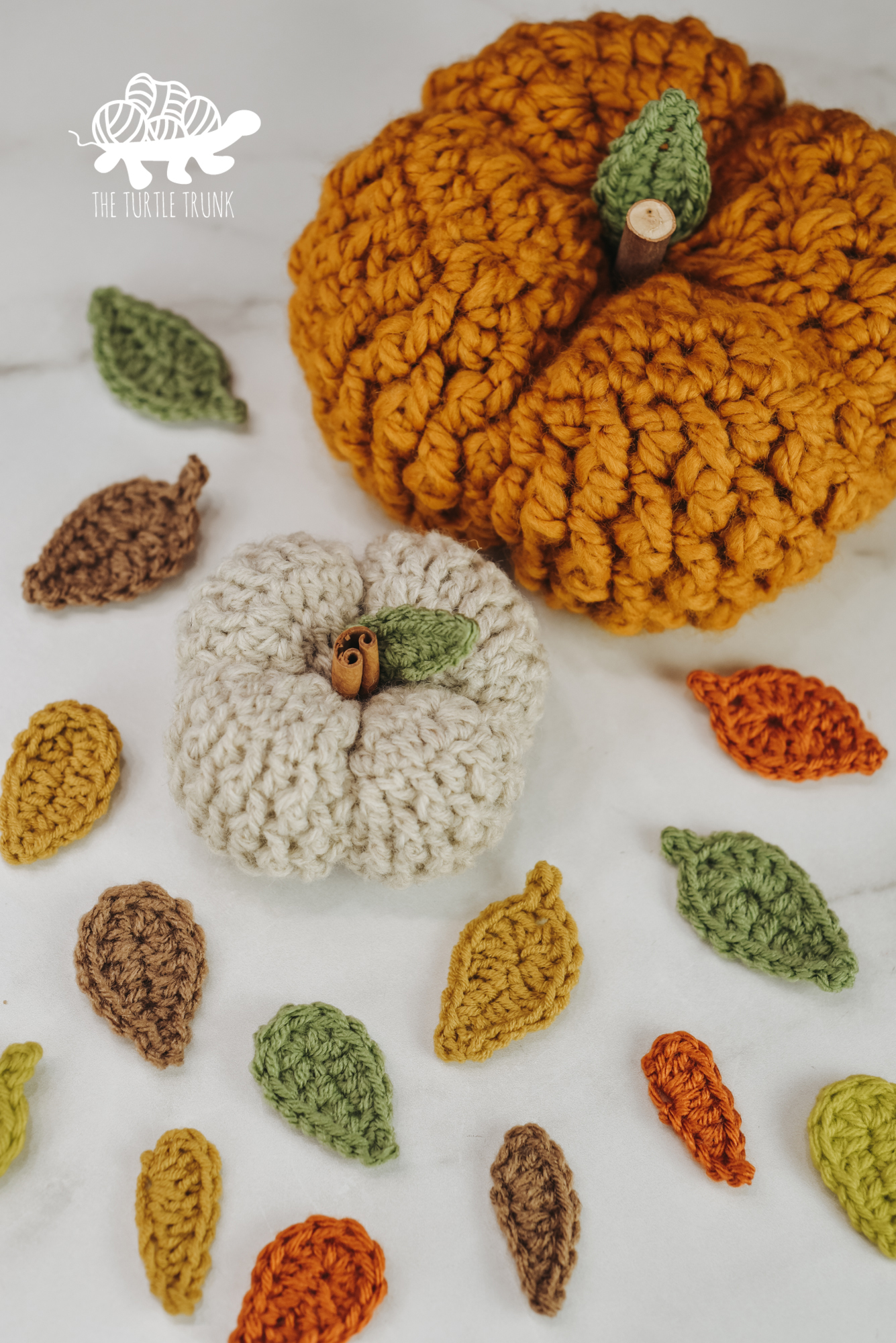 Photo shows 2 crochet pumpkins with crochet leaves surrounding them on a white surface.