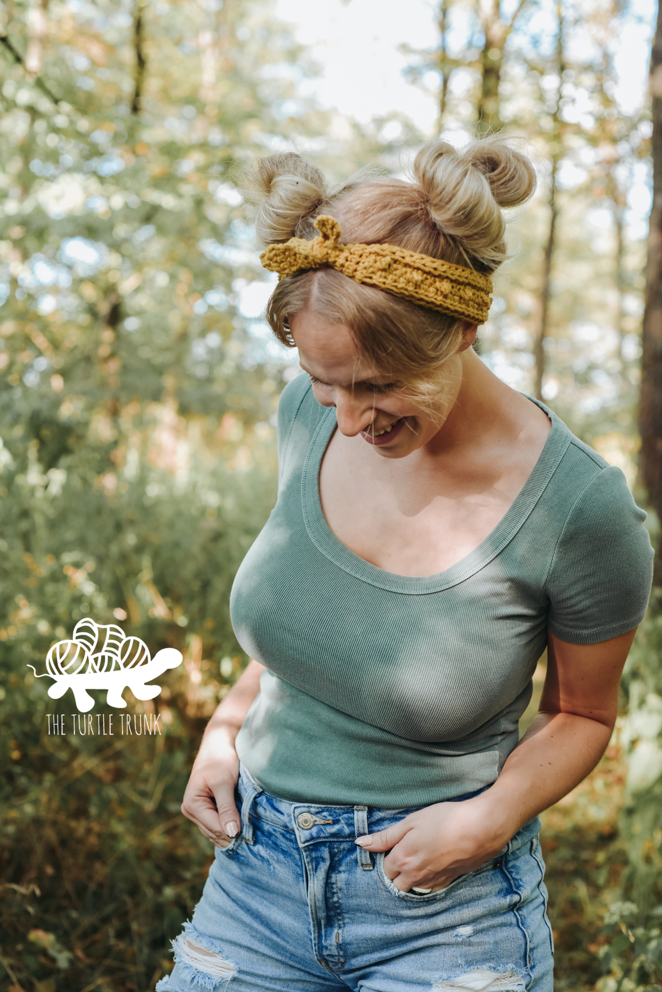 Woman standing outside in the forest wearing a green t-shirt, blue jean shorts, and a yellow, crochet headband. Crochet headband pattern is the Skinny Picot Headband by The Turtle Trunk.