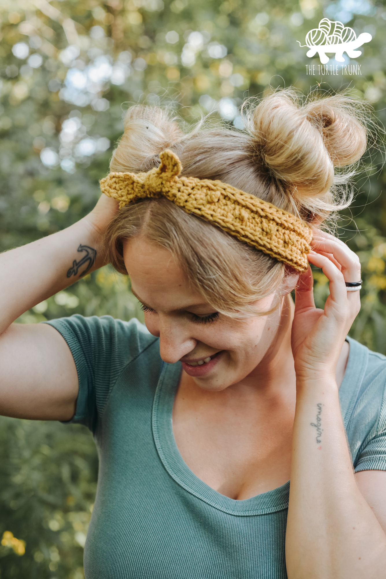 Woman standing outside in the forest wearing a green t-shirt, blue jean shorts, and a yellow, crochet headband. Crochet headband pattern is the Skinny Picot Headband by The Turtle Trunk.