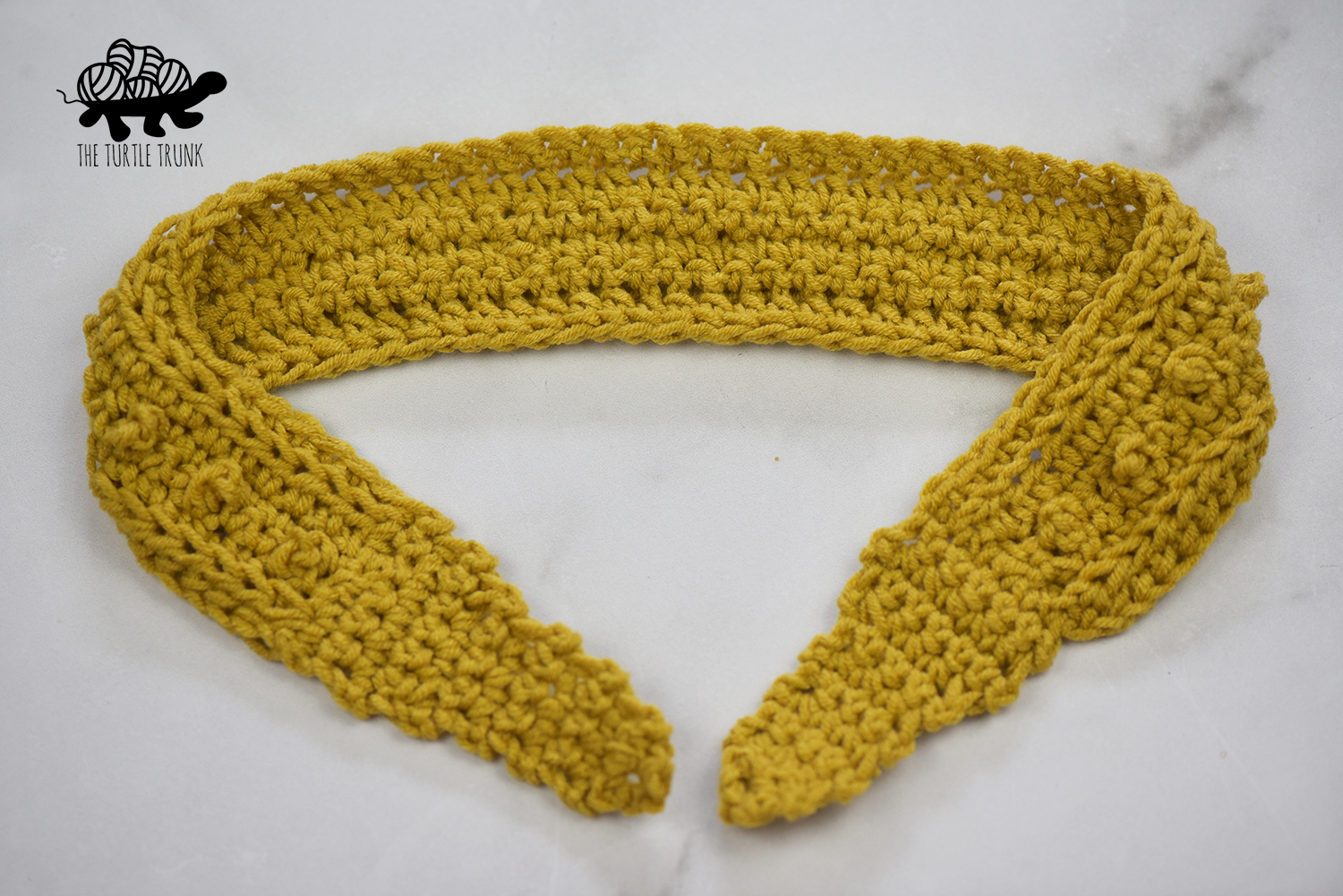 Photo shows a untied, crochet headband laying on a white surface. The crochet headband pattern is the Skinny Picot Headband by The Turtle Trunk.