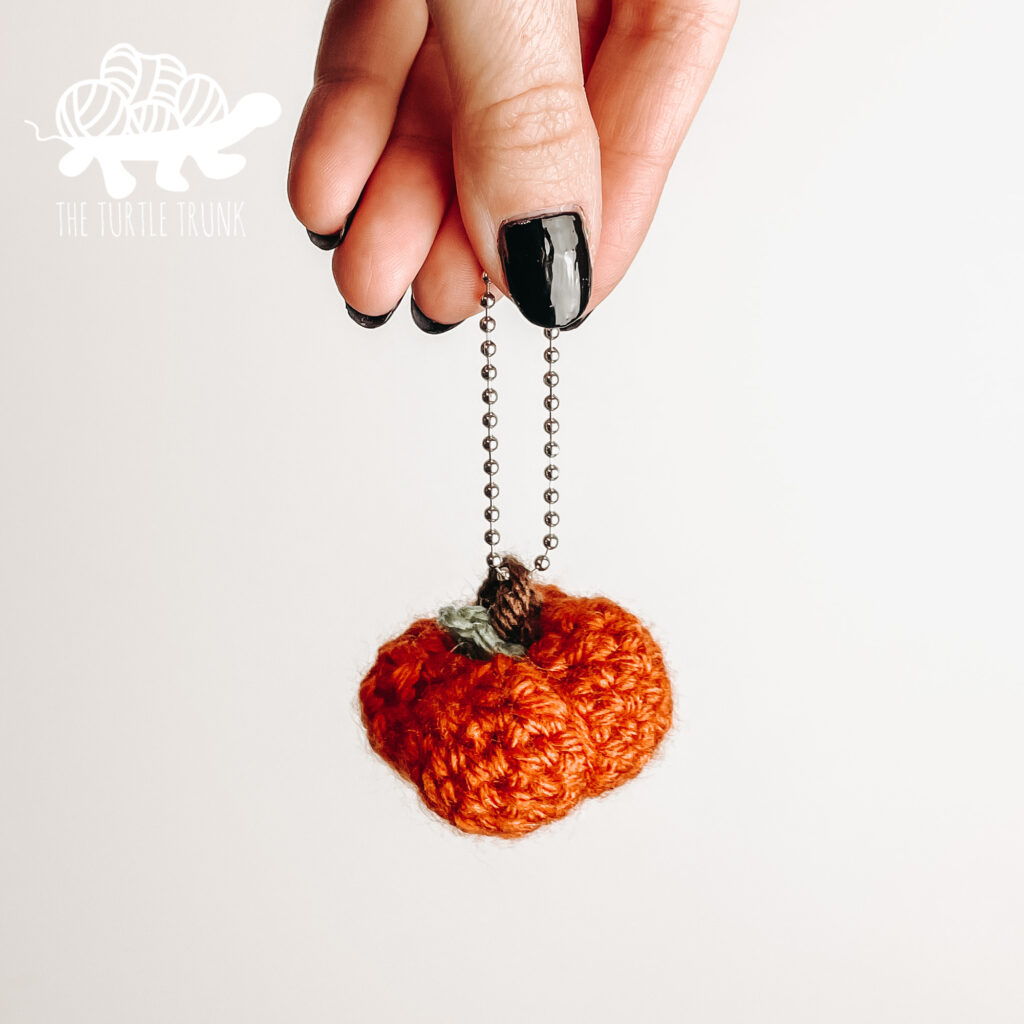 Photo shows a hand holding a mini crochet pumpkin by a chain. Mini Pumpkin crochet pattern by The Turtle Trunk.