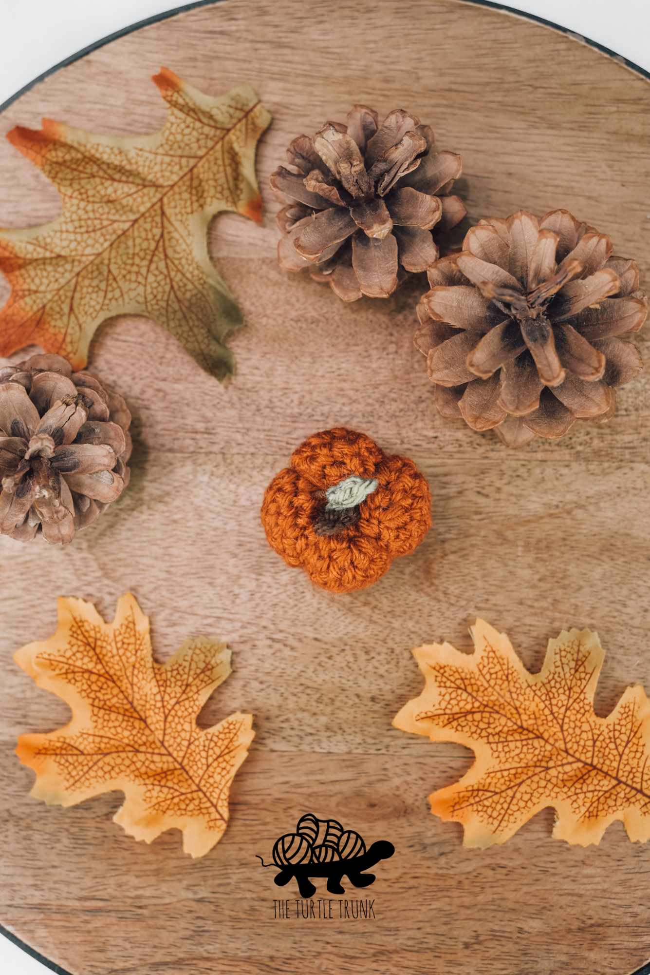 Photo shows a mini crochet pumpkin sitting on a wooden board with pinecones and leaves laying around it. Mini Pumpkin crochet pattern by The Turtle Trunk.