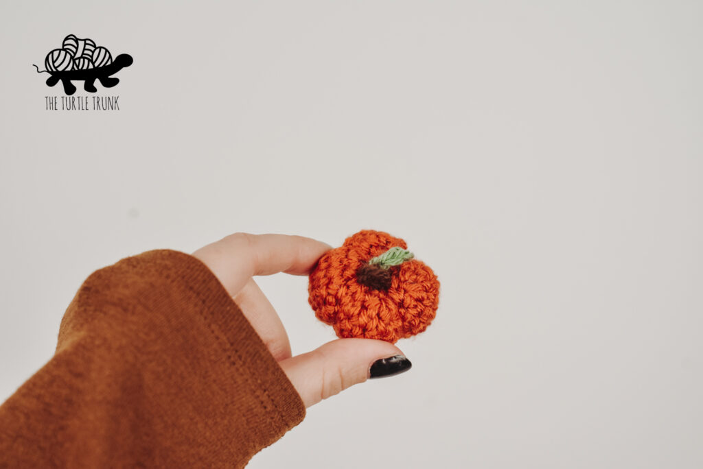 Photo shows a hand holding a mini crochet pumpkin. Pattern by The Turtle Trunk.