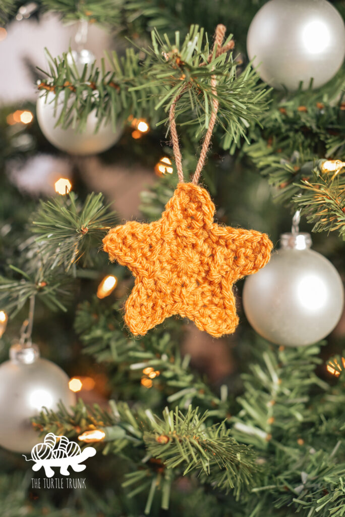 A yellow crochet star is hanging on a Christmas tree as an ornament. Star crochet pattern by The Turtle Trunk.