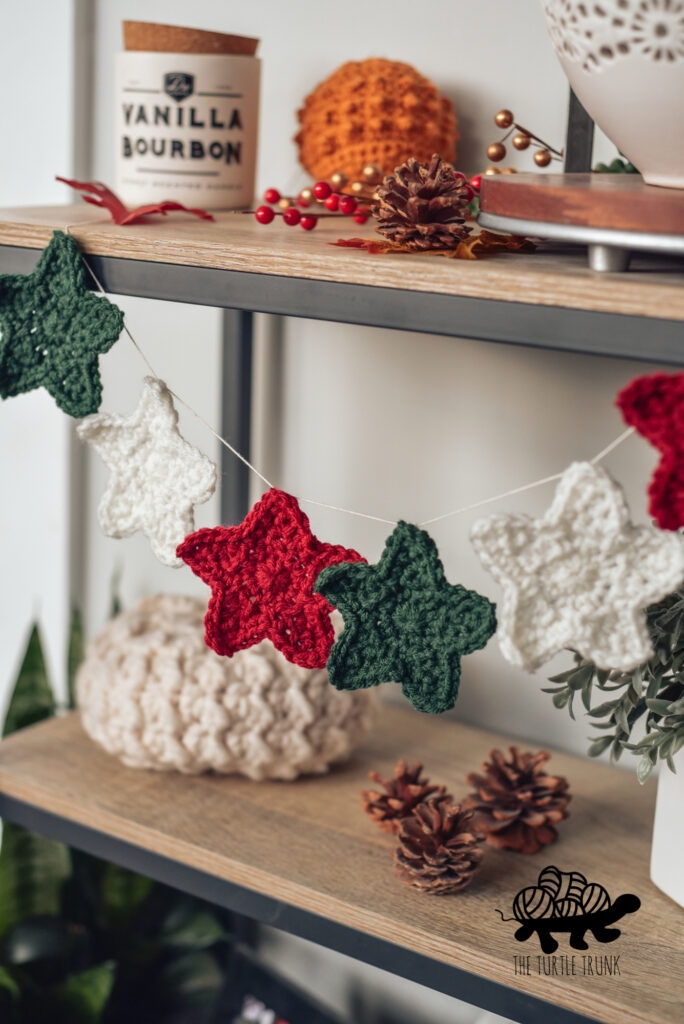 Red, white, and green crochet stars are tied to a string as a garland, hanging on a shelf. Star crochet pattern by The Turtle Trunk.