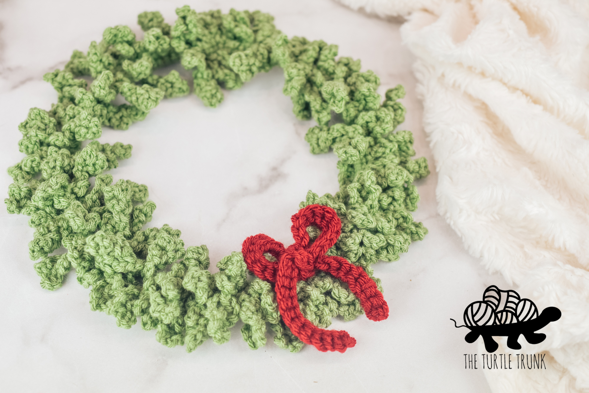 Photo shows a green, crochet wreath with a red bow laying on a white surface. Crochet pattern is the Happy Holiday Wreath by The Turtle Trunk.