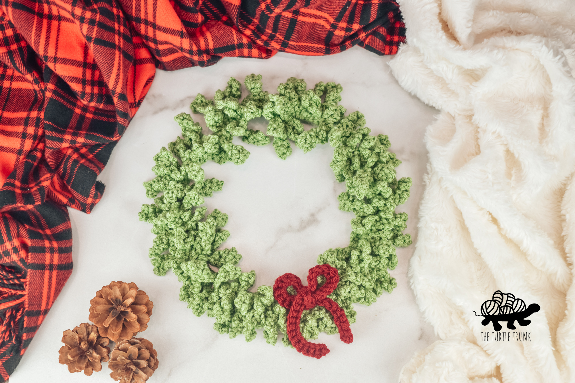 Photo shows a green, crochet wreath with a red bow, laying on a white surface surrounded by blankets. Crochet pattern is the Happy Holiday Wreath by The Turtle Trunk.