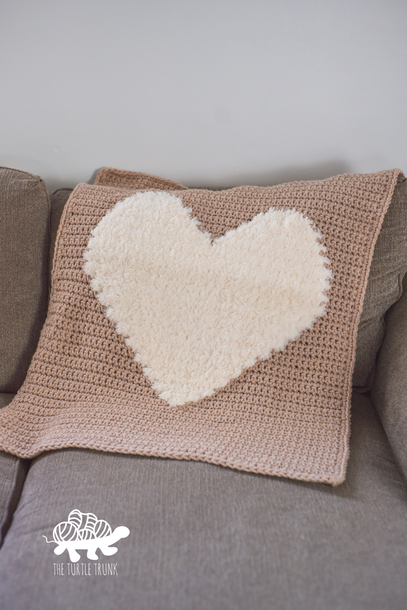 Photo shows a blush pink, crochet blanket with a white faux fur heart on it, laying on a couch. The crochet pattern is the Love to Cuddle Blanket by The Turtle Trunk.