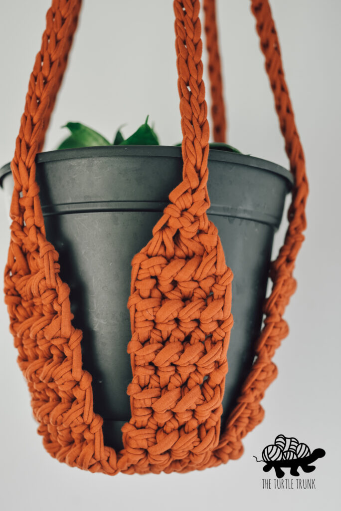 Photo shows an orange, crochet plant hanger with a black plant pot in it, hanging from the ceiling.