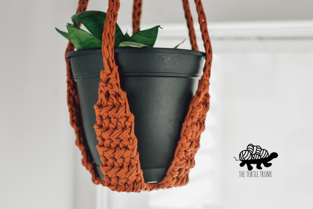 Photo shows a crochet plant hanger holding a black potted plant and hanging from the ceiling.