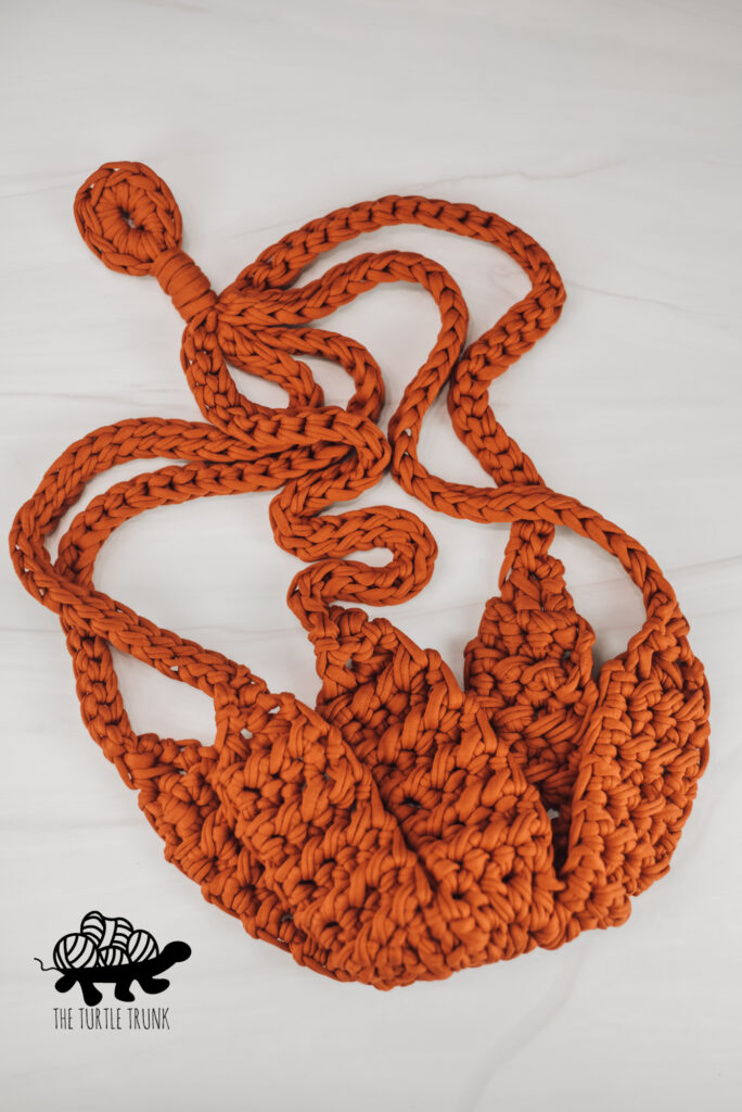 Photo shows an orange, crochet plant hanger laying on a white surface.