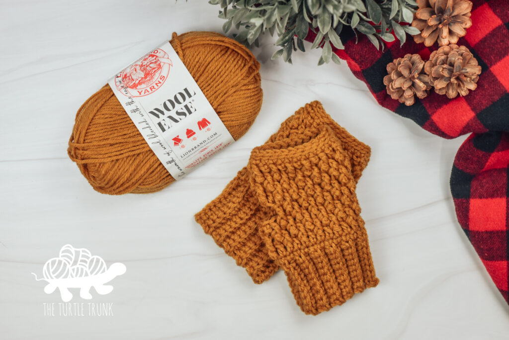 Photo shows a skein of yellow Lion Brand Yarn sitting beside a pair of crochet fingerless gloves on a white surface.