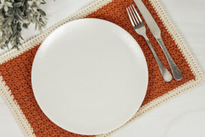 A orange and white crochet placemat on a white surface with a white plate on top of it.