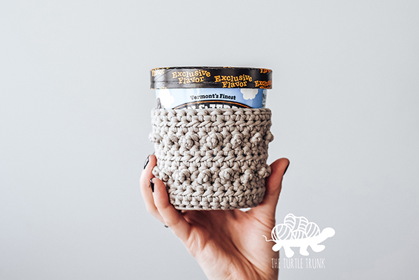 Hand holding a pint of ice cream with a crochet cozy on it.