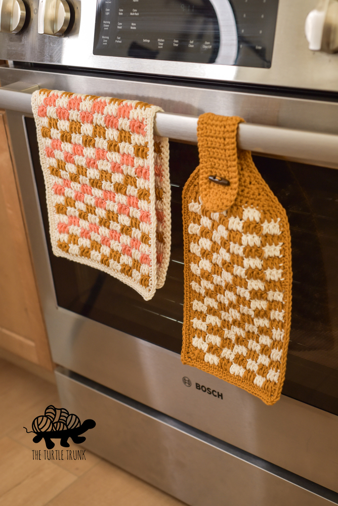 A crochet checkered dish towel and hanging towel on a stove.