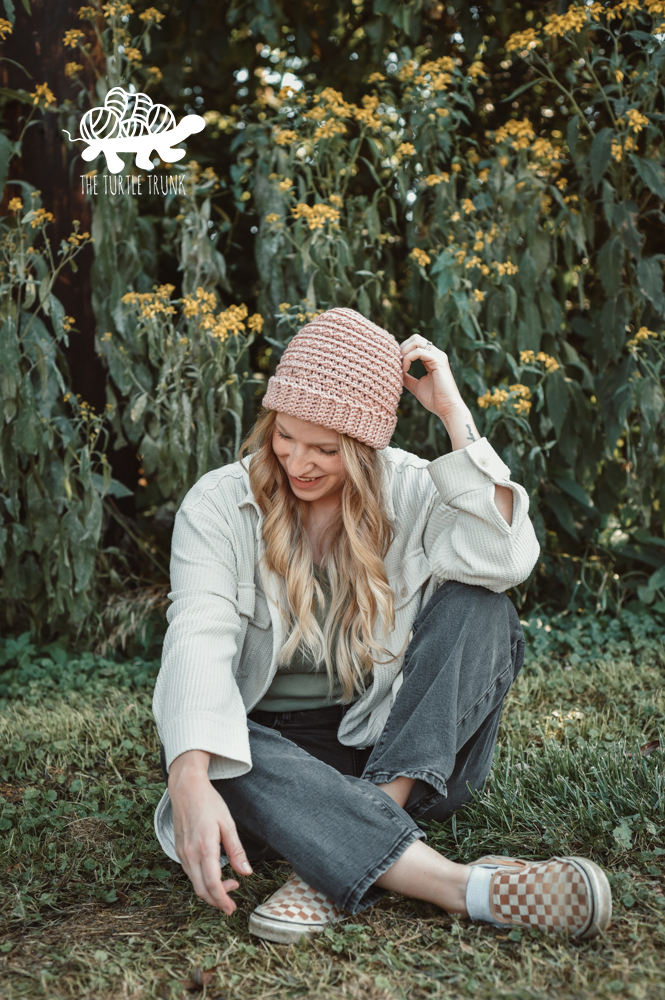 Woman sitting in the grass, wearing a pink, crochet beanie.