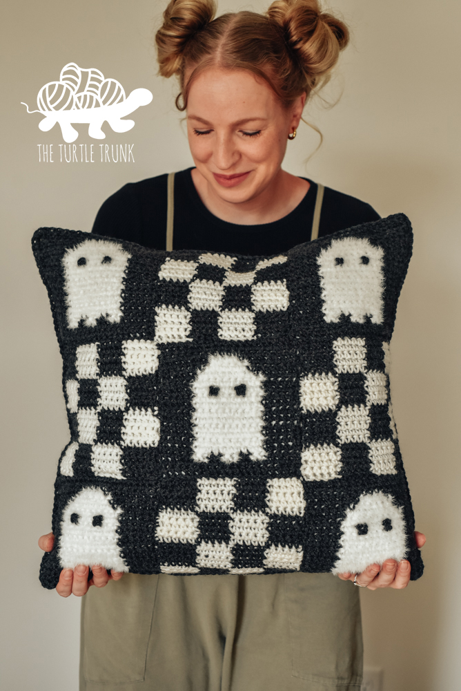 Woman holding a crochet pillow with a checkered pattern with ghosts on it.