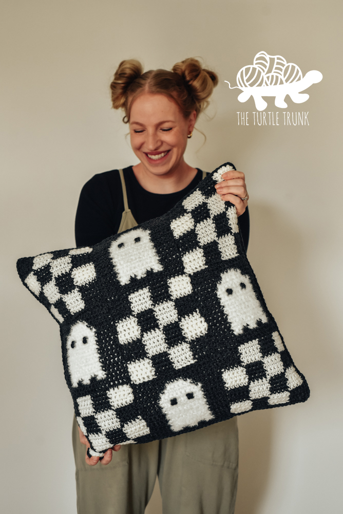 Woman holding a crochet pillow with a checkered pattern with ghosts on it.