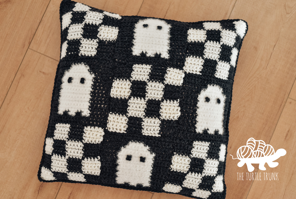 A black and white, crochet pillow with a checkered pattern and ghosts on it.