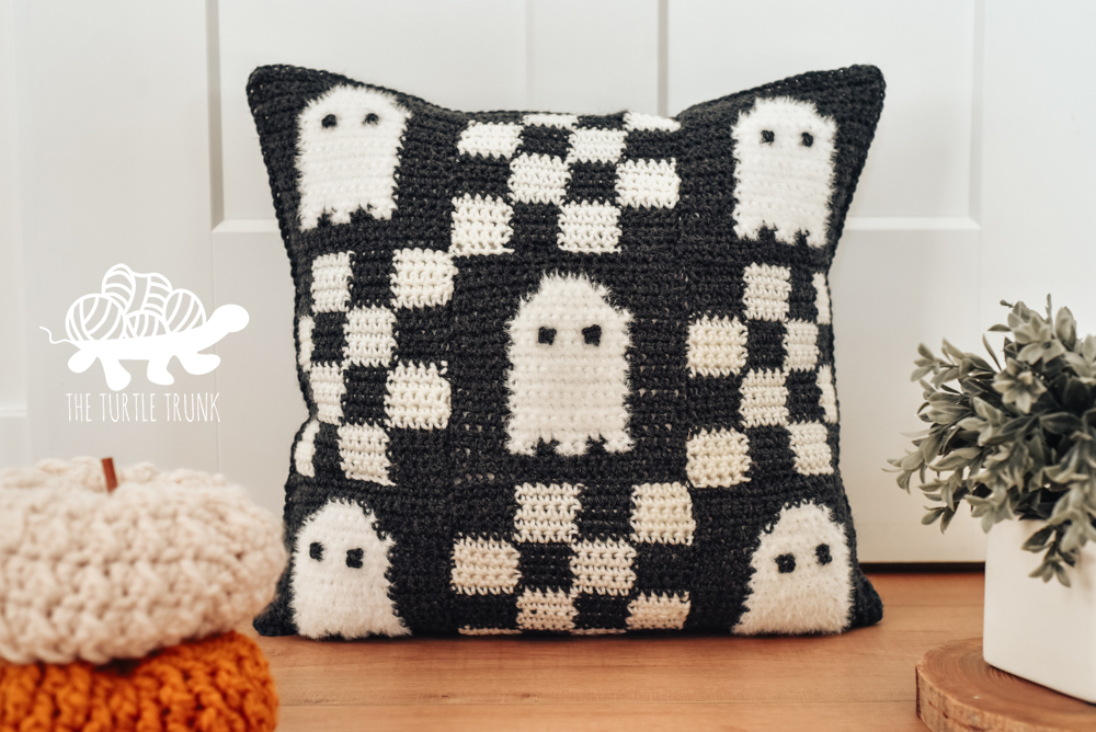 A black and white crochet pillow with a checkered pattern and ghosts on it.