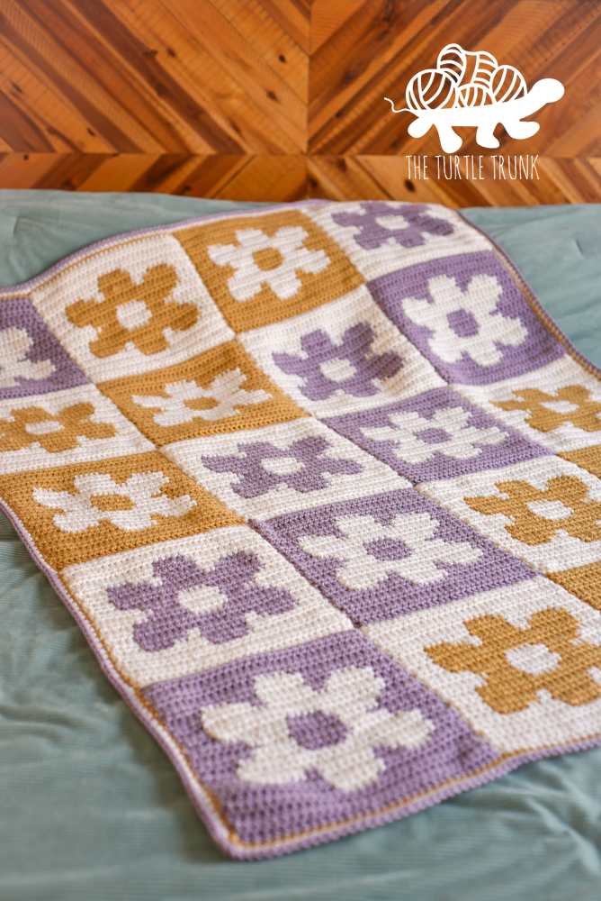 A yellow and purple, checkered flower crochet blanket laying on a bed.